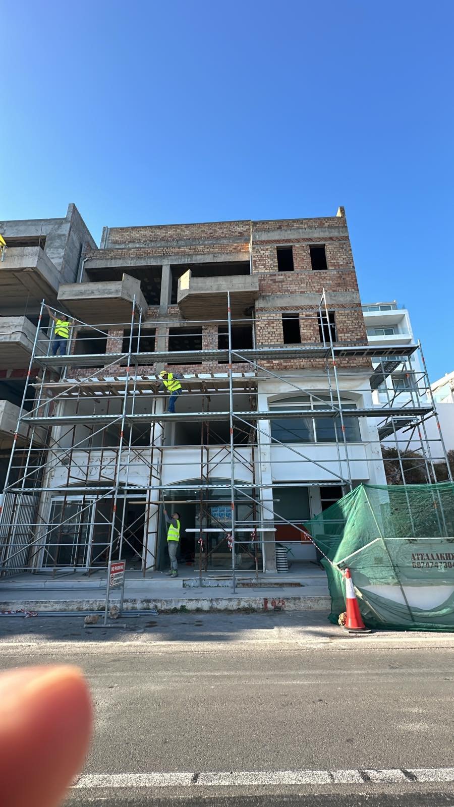 A multi-story building under construction with scaffolding, construction workers in yellow vests working on the exterior, and a clear blue sky overhead.