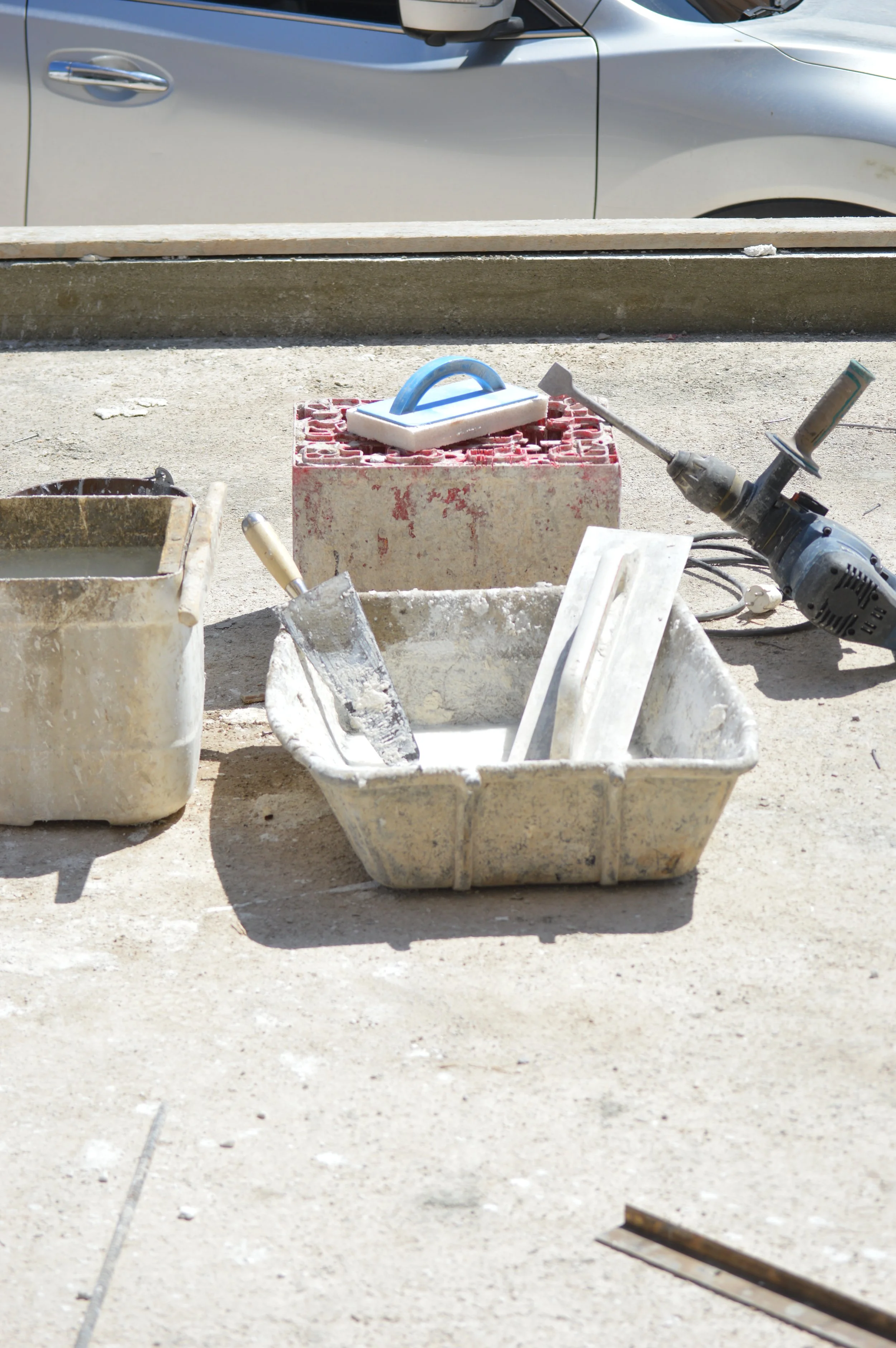 Construction site with buckets of wet cement, a trowel, a float, and a portable power drill, with a car in the background.