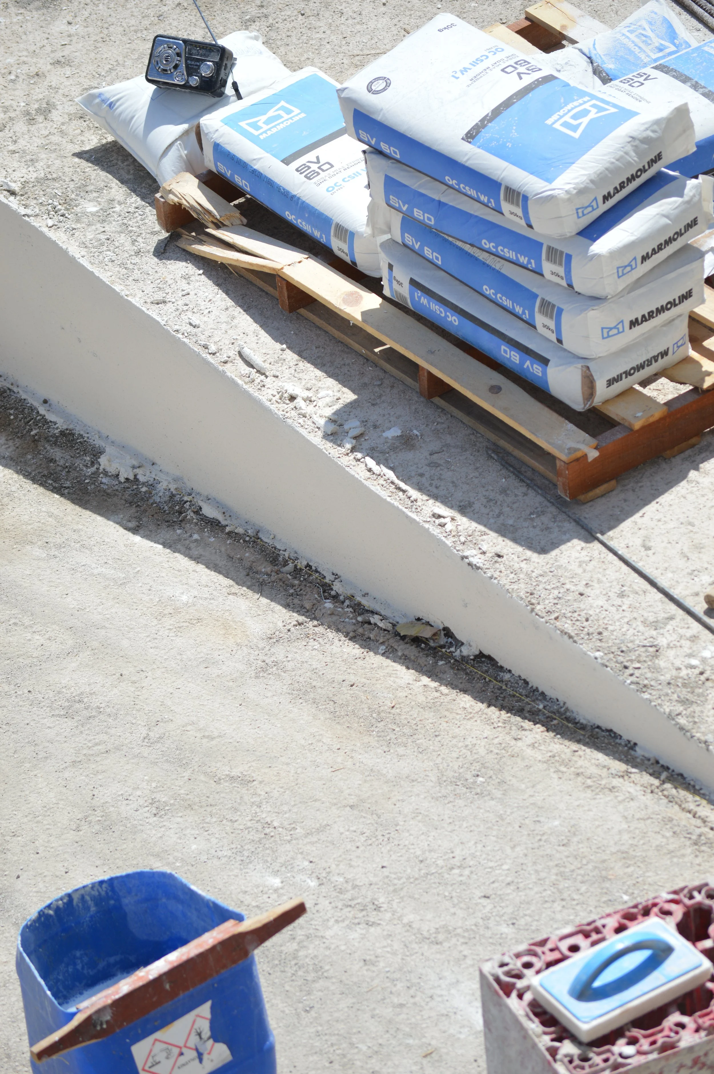 Construction site with bags of cement, a tape measure, a blue bucket, and a stack of bricks.