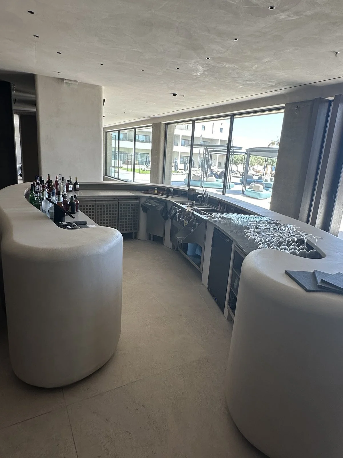 Empty modern bar with liquor bottles, glasses, and view of outdoor pool area through large windows.