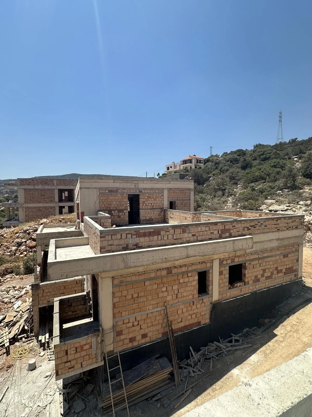 A multi-story house under construction with brick walls, concrete beams, and open window and door spaces, set against a hillside with other houses and a clear blue sky.