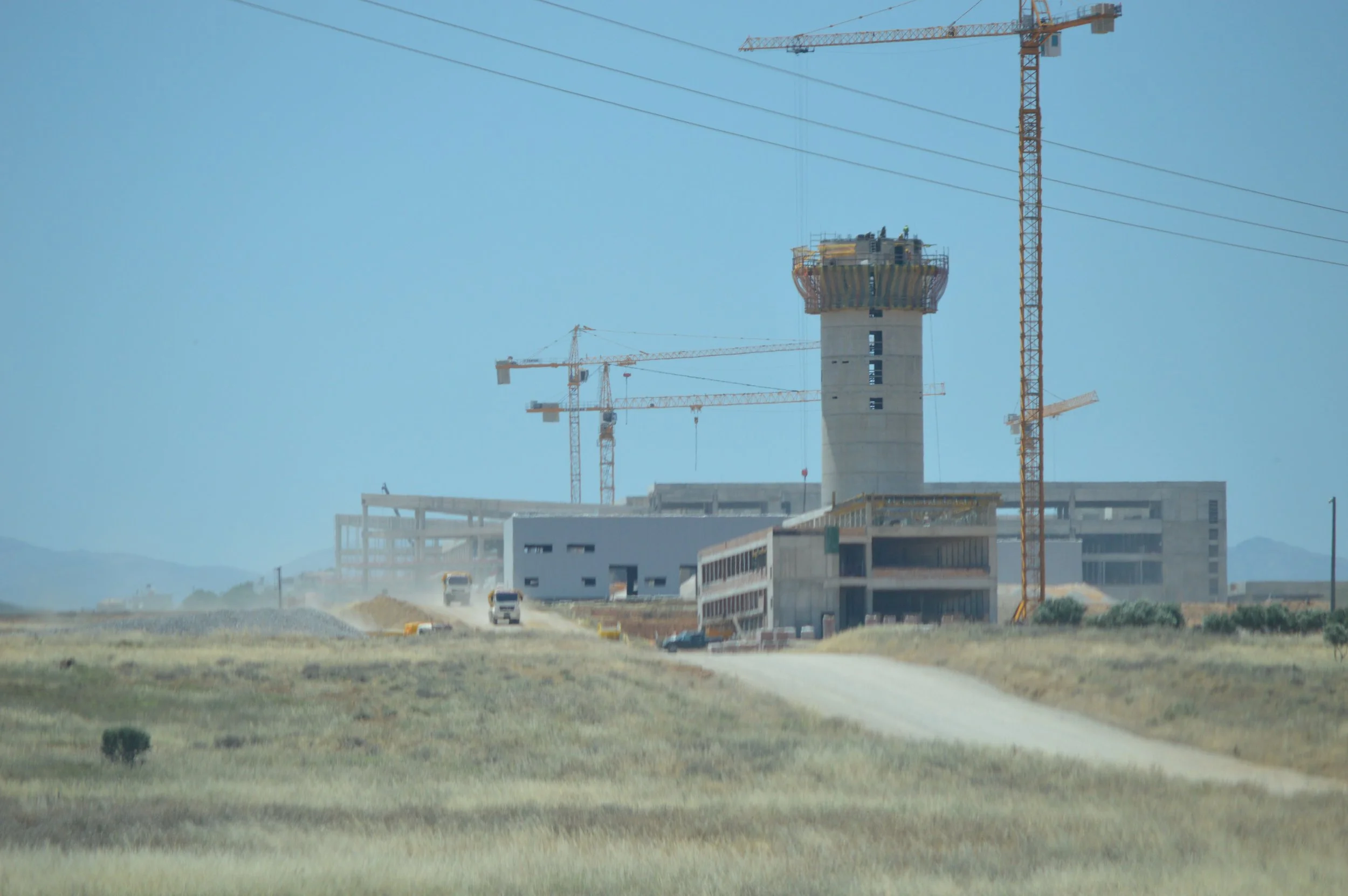 Construction site with multiple cranes and a partially built tall structure, set in a dusty, rural landscape.