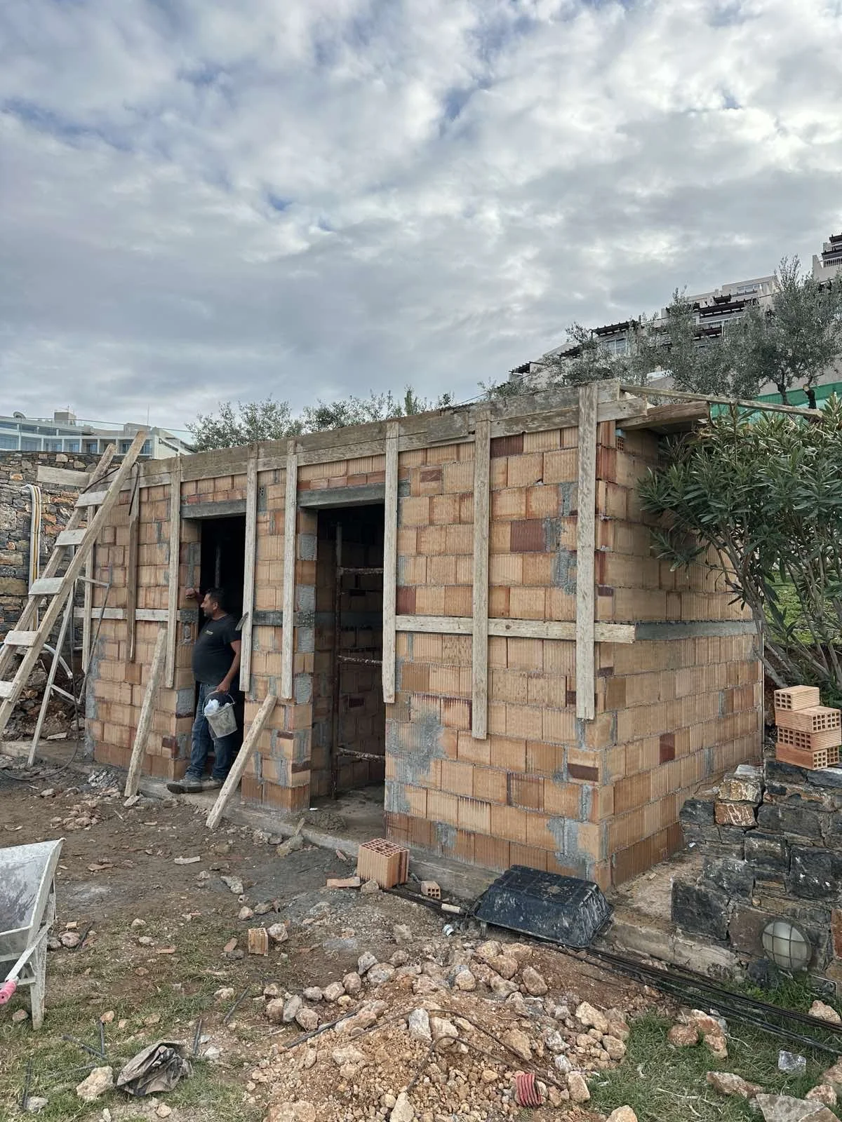 A small brick building under construction with wooden beams and a person standing outside, holding construction tools, on a construction site with scattered rocks and bricks.