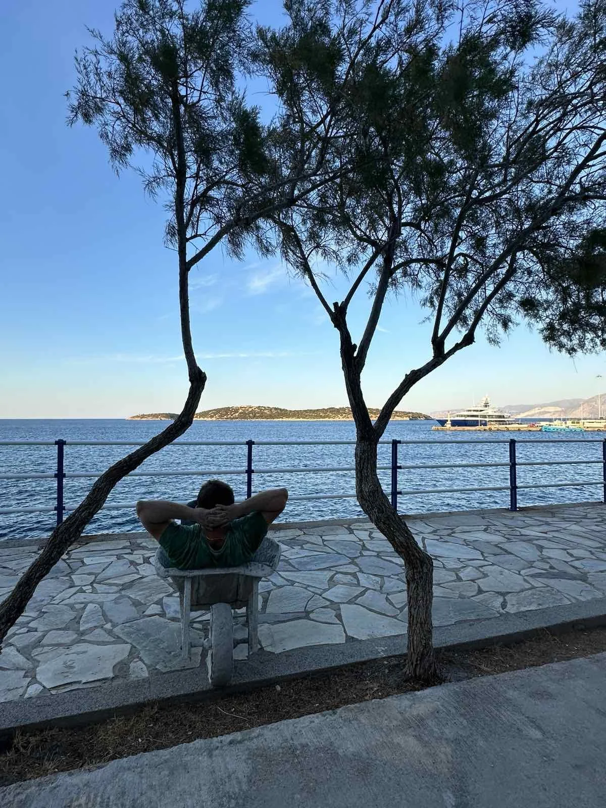 A person sitting on a bench by the water, leaning back with hands behind head under trees, with a view of boats and an island in the distance.