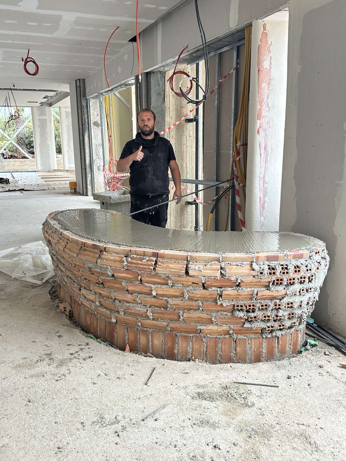 Construction worker standing next to an unfinished brick bar with freshly poured concrete, giving a thumbs up inside a building under renovation.