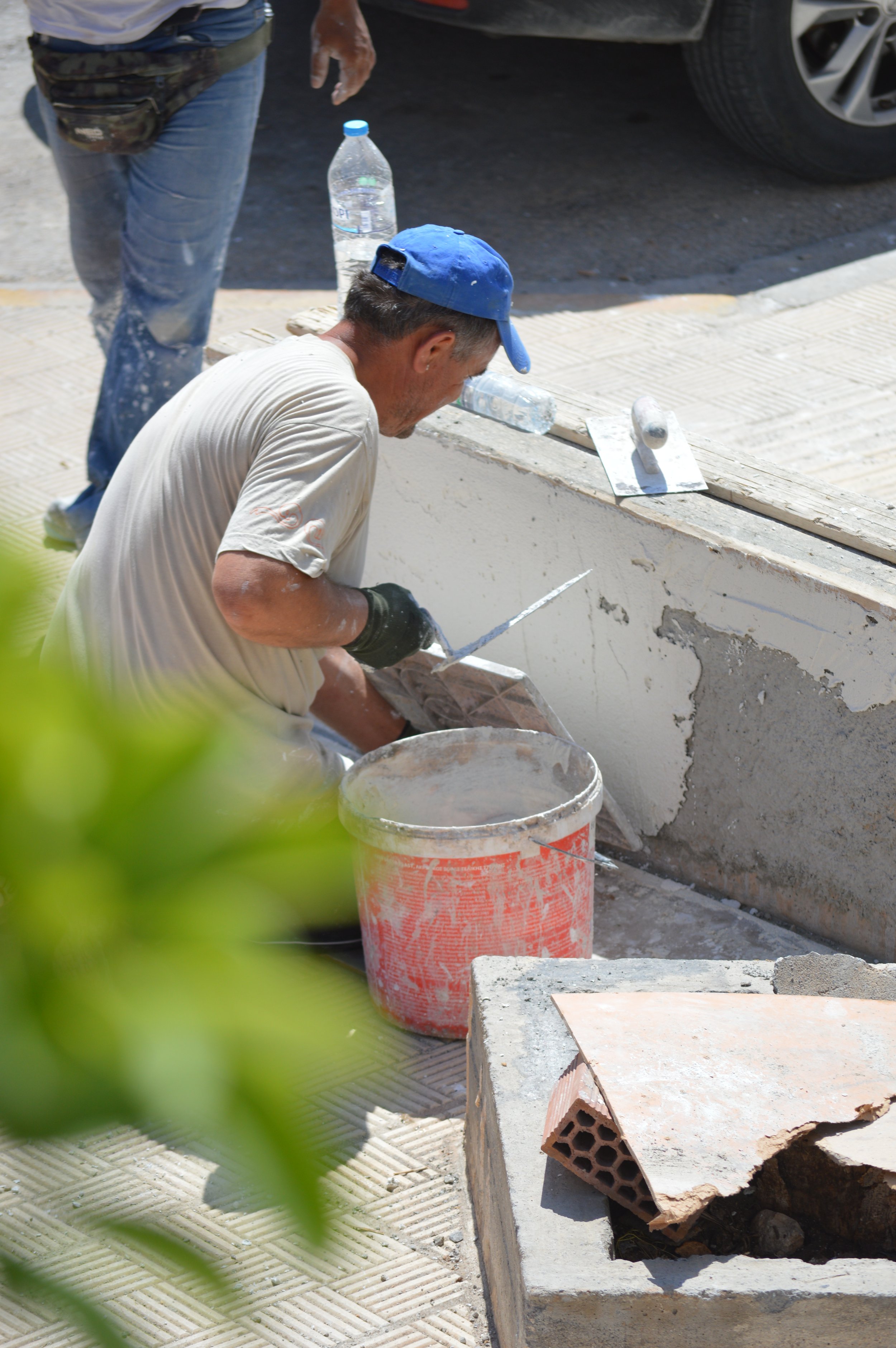 A man wearing a blue cap and gloves is working on a construction site, using a trowel to apply cement or plaster to a wall. There is a bucket of mix, a trowel, and construction materials nearby.