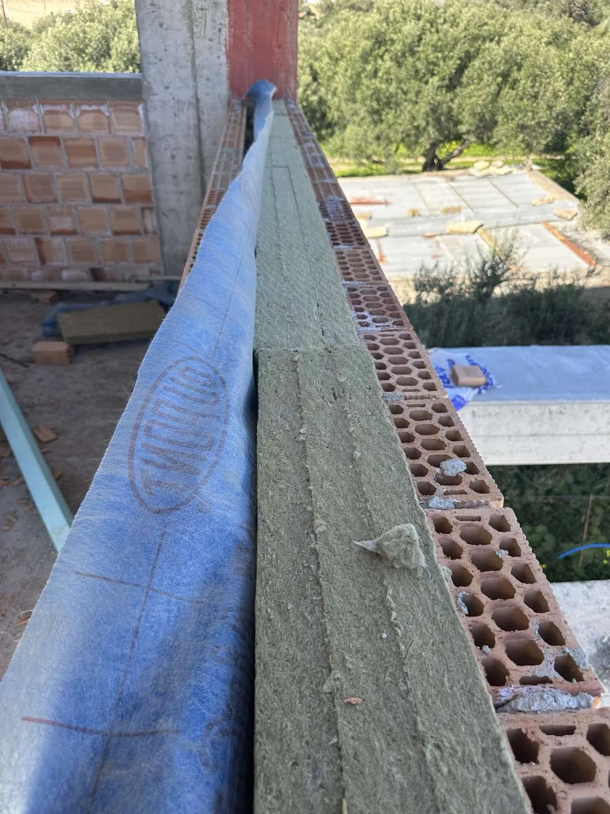 A construction site showing a brick wall and insulation material on a balcony or ledge. In the background are trees and rooftops of other buildings.