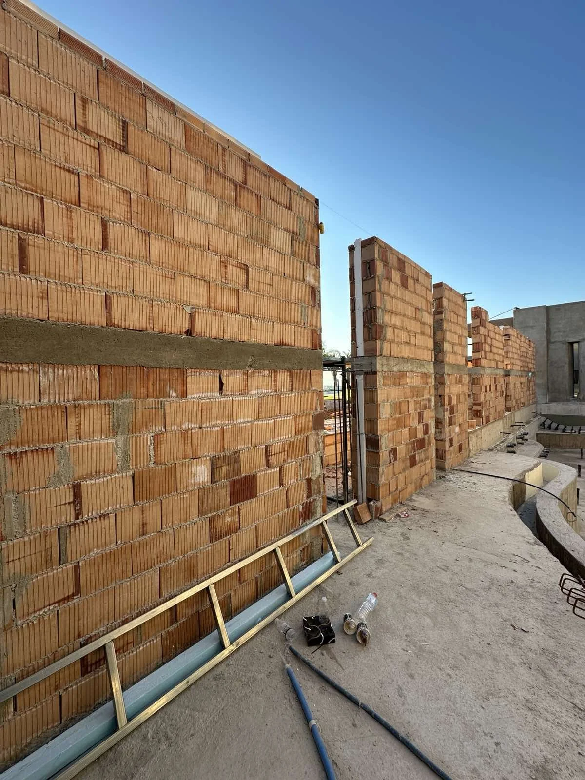 Construction site with brick walls, some partially built, scaffolding, tools, and water bottles on the ground under a clear blue sky.