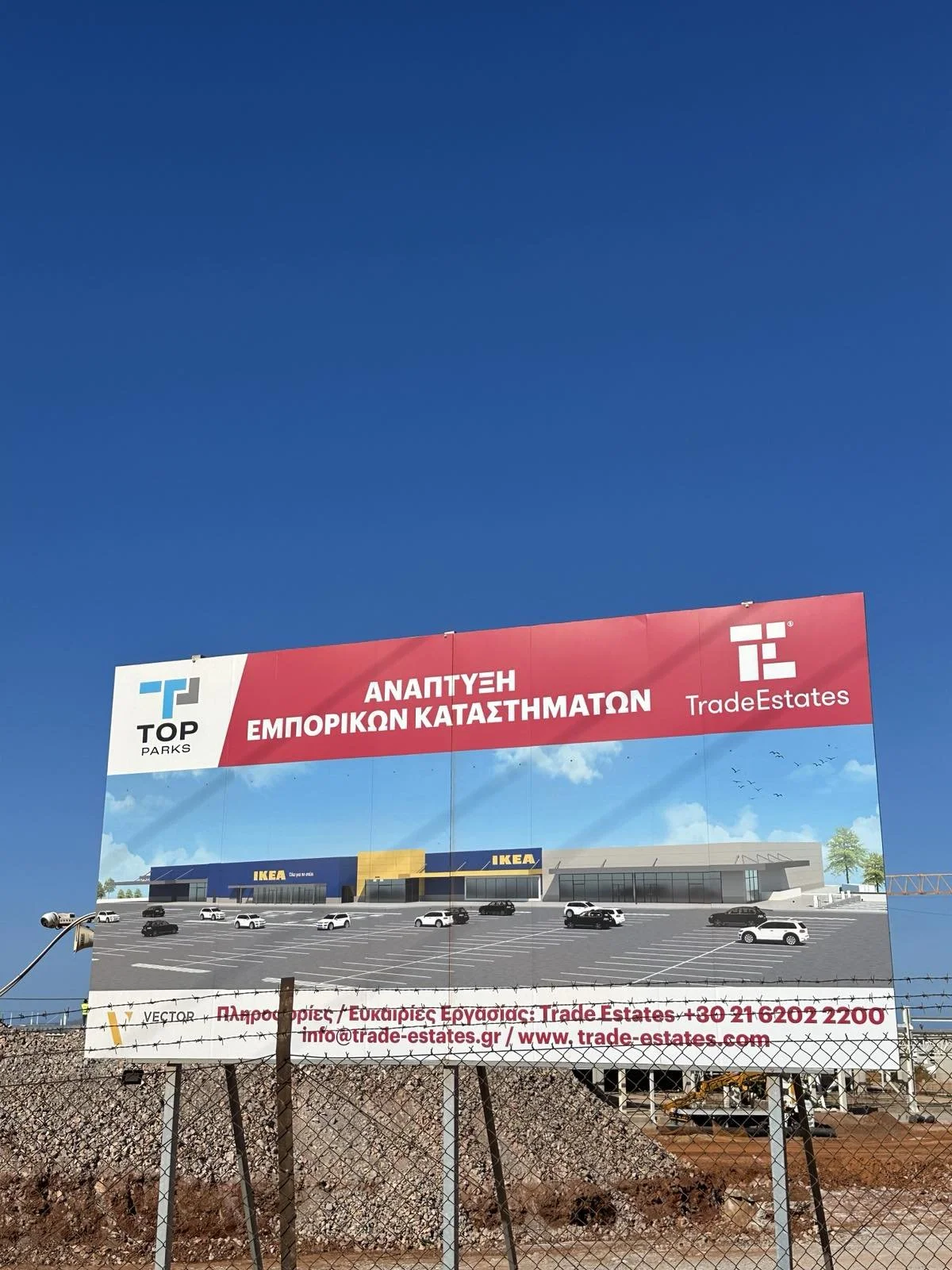 A billboard advertising the development of commercial stores with a rendering of a shopping center, including IKEA, against a bright blue sky. The text is in Greek and English, mentioning trade estates and contact information.