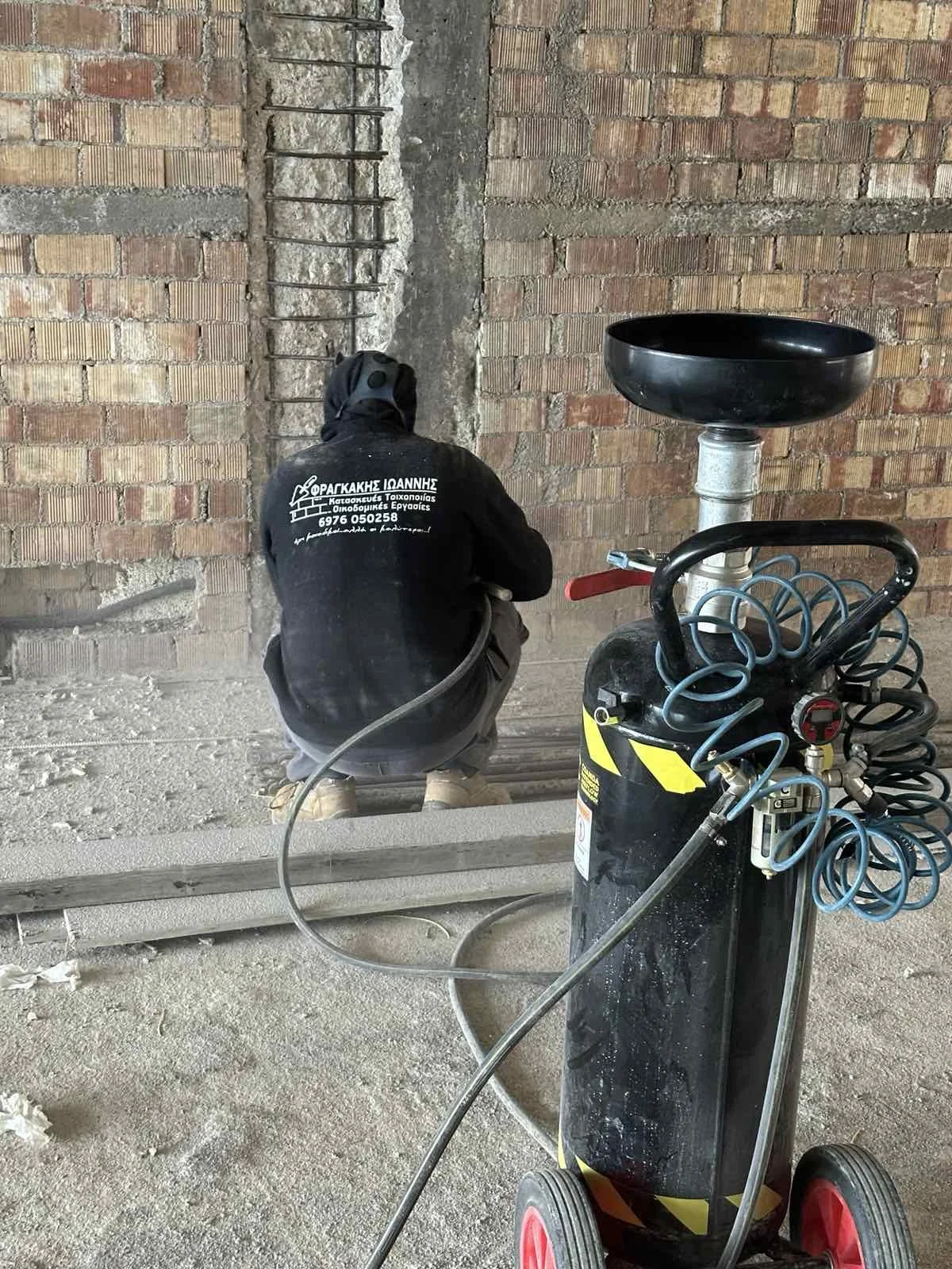 A construction worker crouching and working on a brick wall with exposed rebar, surrounded by construction tools and equipment, in an unfinished building.
