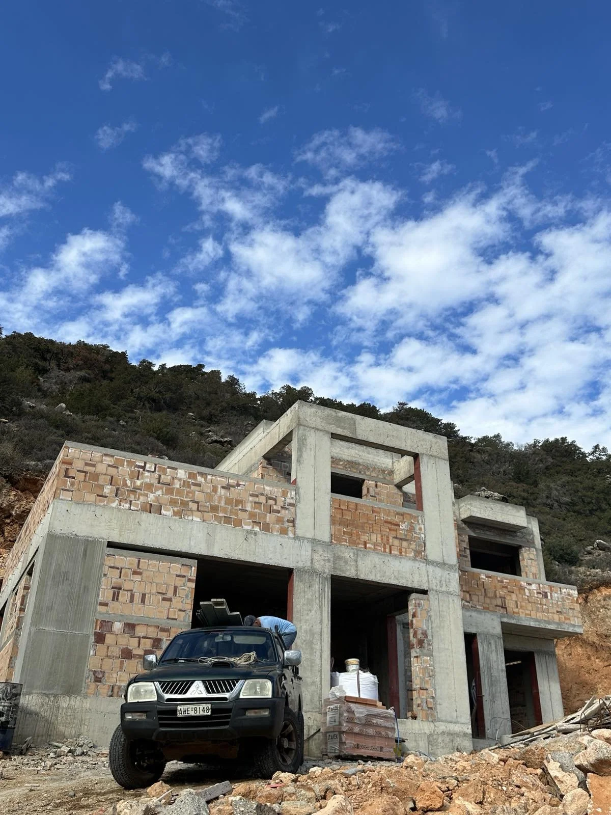 Under construction house with brick and concrete walls, parked black SUV in front, construction materials nearby, hillside in background, blue sky with clouds above.