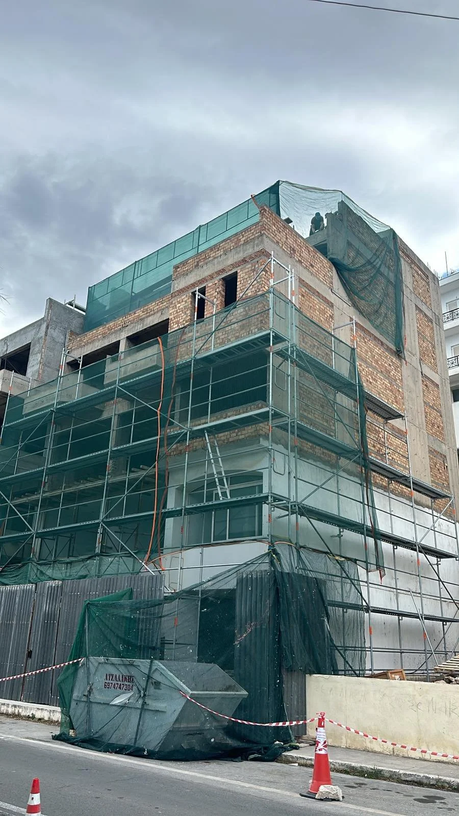 Construction site of a multi-story building with scaffolding, green safety netting, and workers on the top floor, with a cloudy sky in the background.