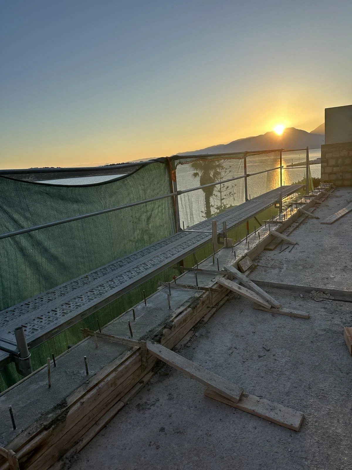 Construction site on a balcony or roof with a view of a body of water, mountains, and a setting sun, with construction materials and protective green netting.