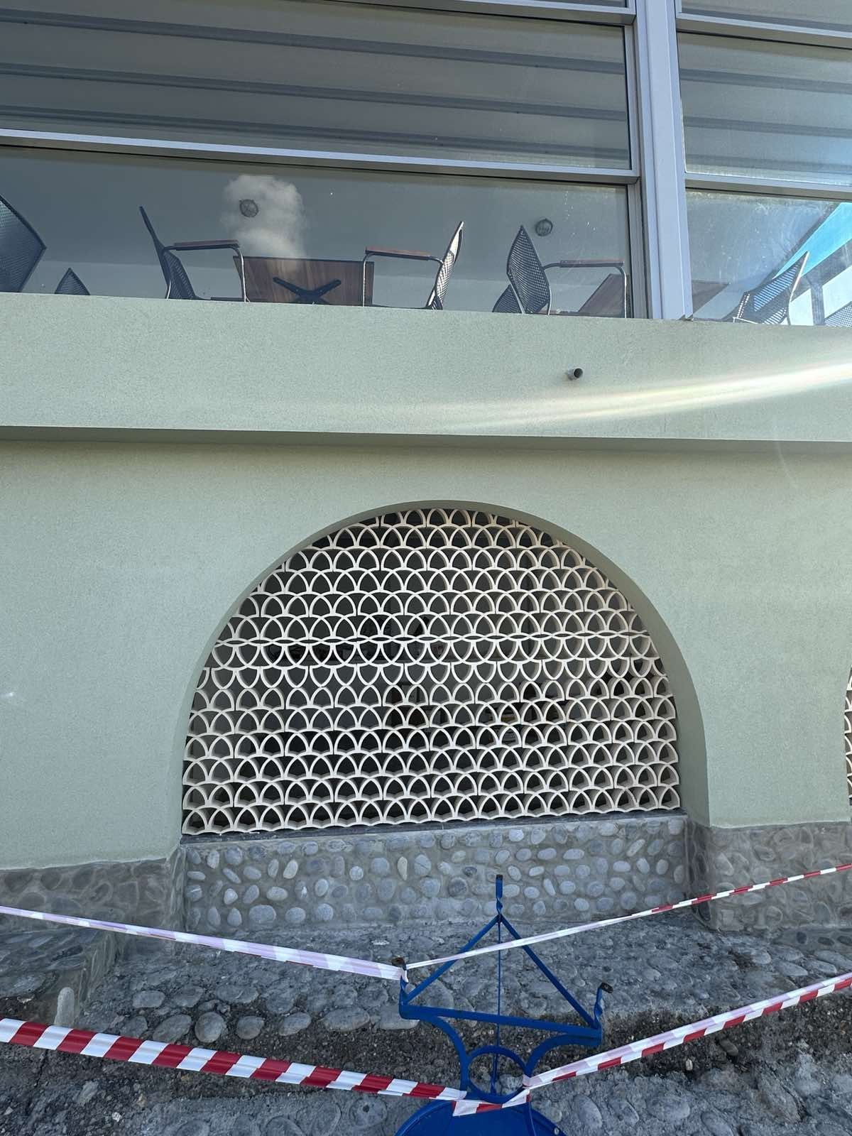Outside view of a building with a decorative arch vent, mesh-covered opening, light green wall, and a gravel area with red and white caution tape and a blue foldable chair.