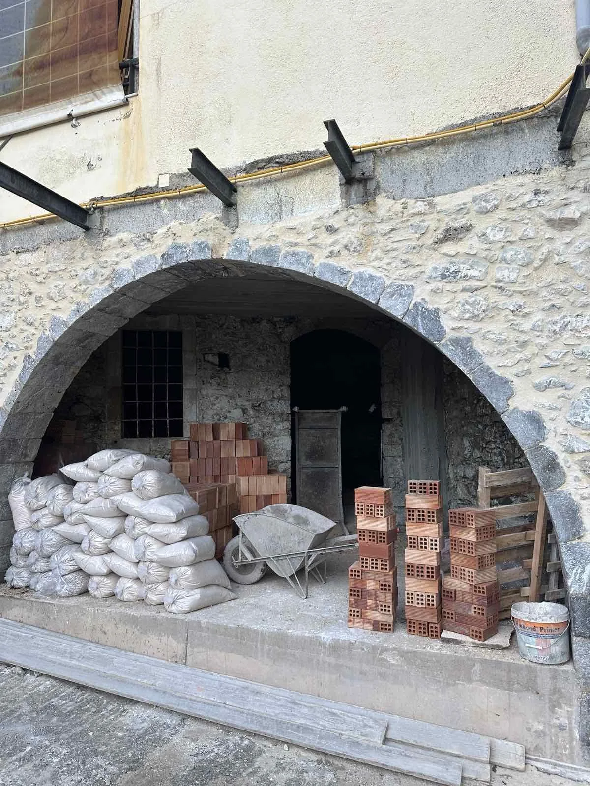 Construction site with bags of concrete, stacks of bricks, a wheelbarrow, and construction tools under an arched stone structure.