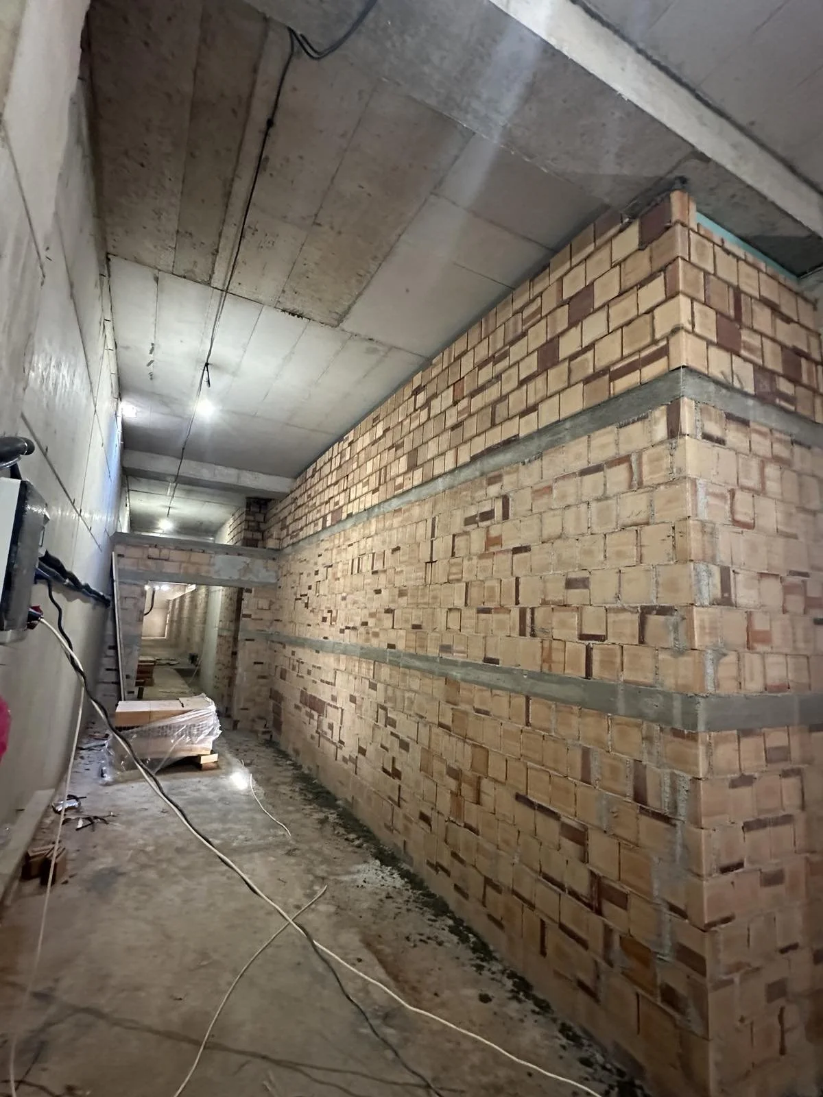 Construction site with brick and concrete walls and ceiling, electrical wires hanging, and construction materials on the floor.
