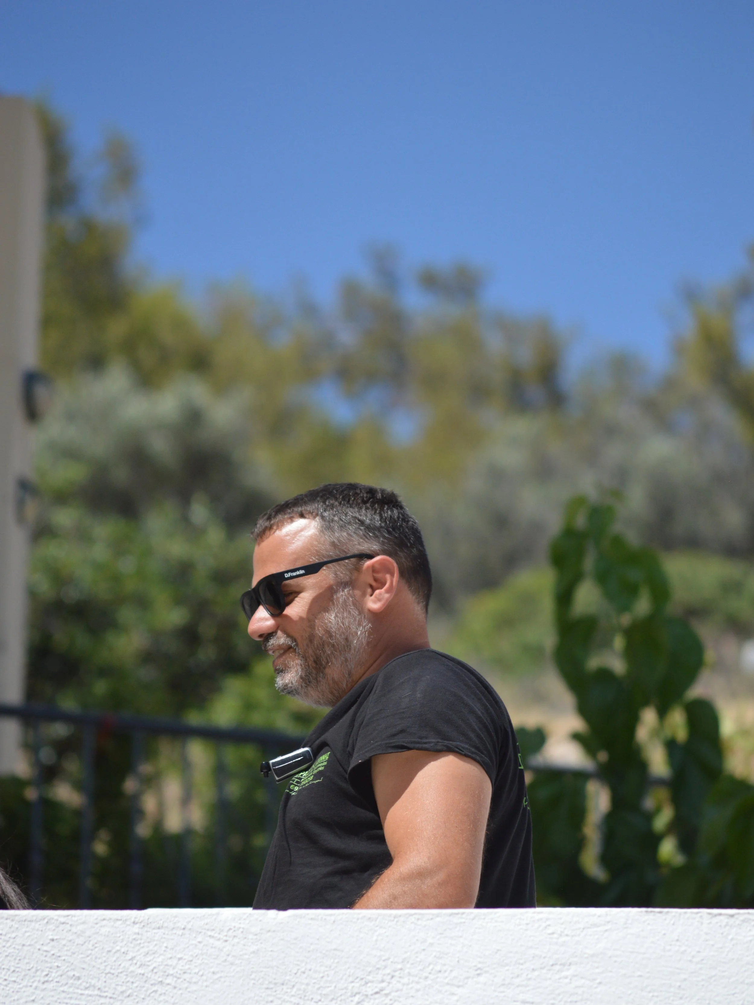 A man with sunglasses and a beard, wearing a black T-shirt, walking outdoors on a sunny day with a blue sky and green trees in the background.