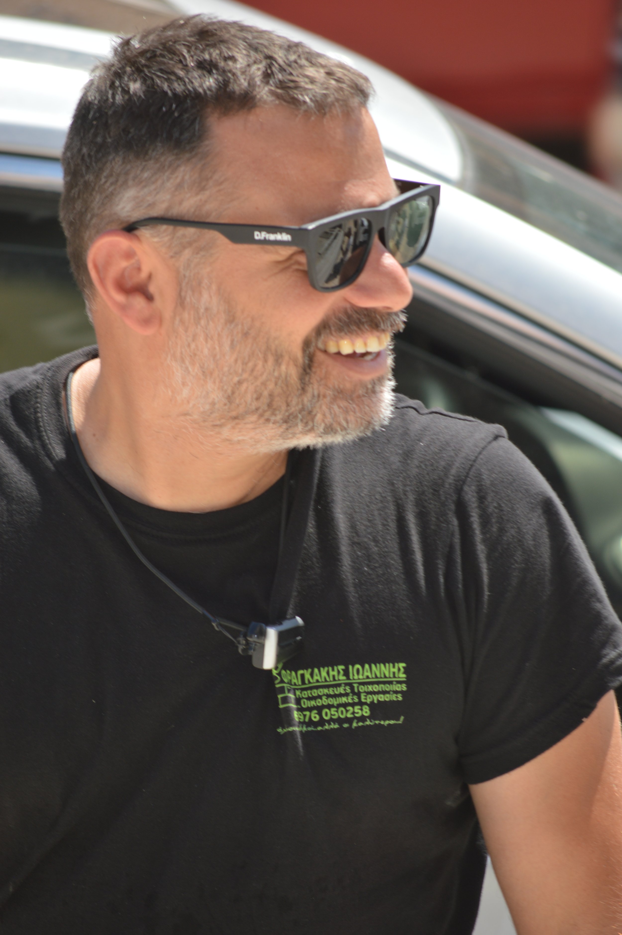 A man with short hair, sunglasses, and a beard, smiling and wearing a black t-shirt with Greek writing, outdoors near a vehicle.