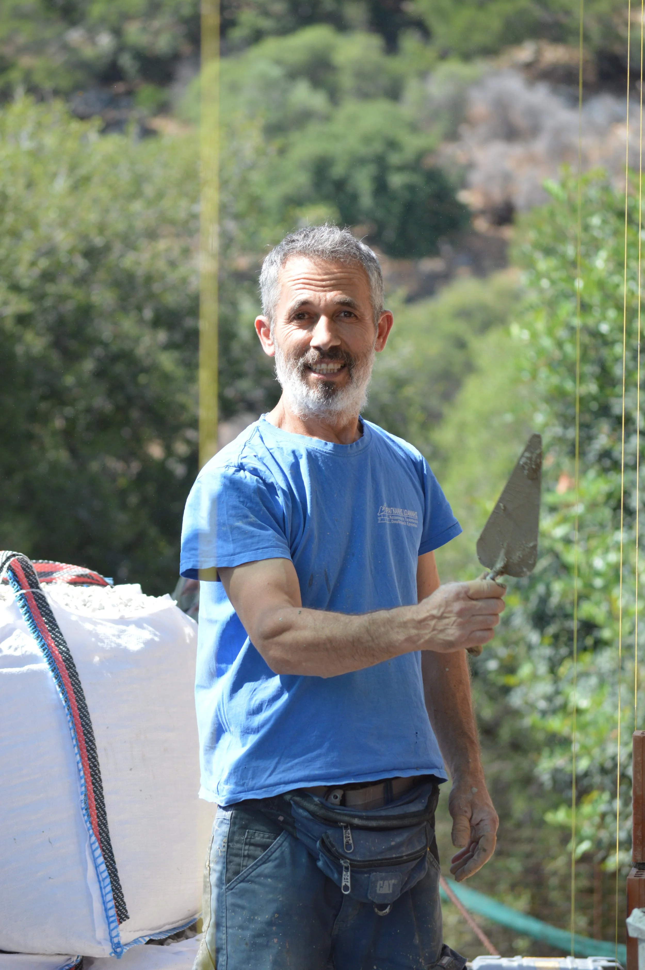 A smiling man with gray hair and beard, wearing a blue T-shirt and work pants, holding a trowel, standing outdoors with greenery in the background.
