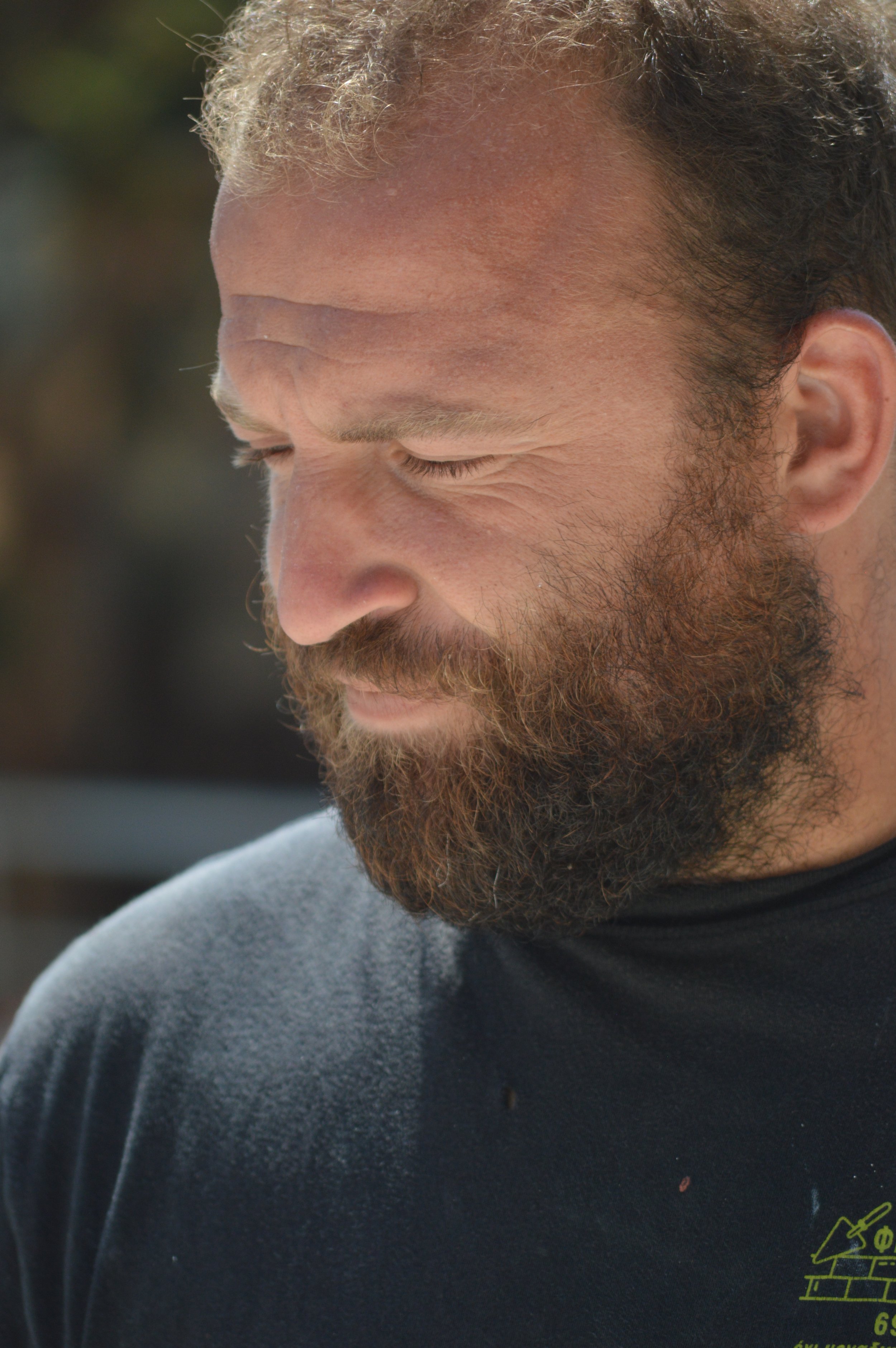 Close-up of a man with a full beard and curly hair, looking down, wearing a black shirt, outdoors.
