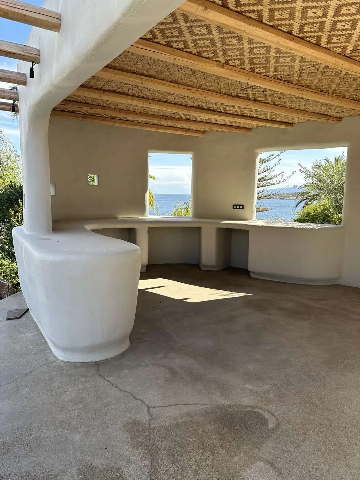 An outdoor space with a built-in white stucco bar area, large open window views of the ocean, and tropical trees, with a wooden ceiling and concrete floor.