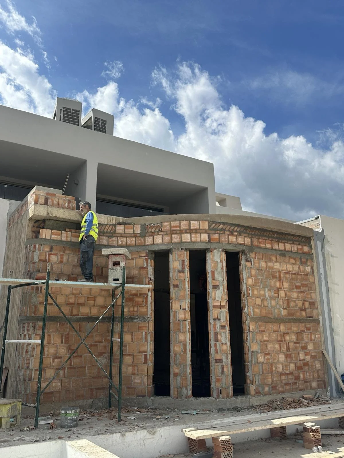 A construction worker standing on scaffolding at a brick building under construction, with a modern white building and blue sky with clouds in the background.