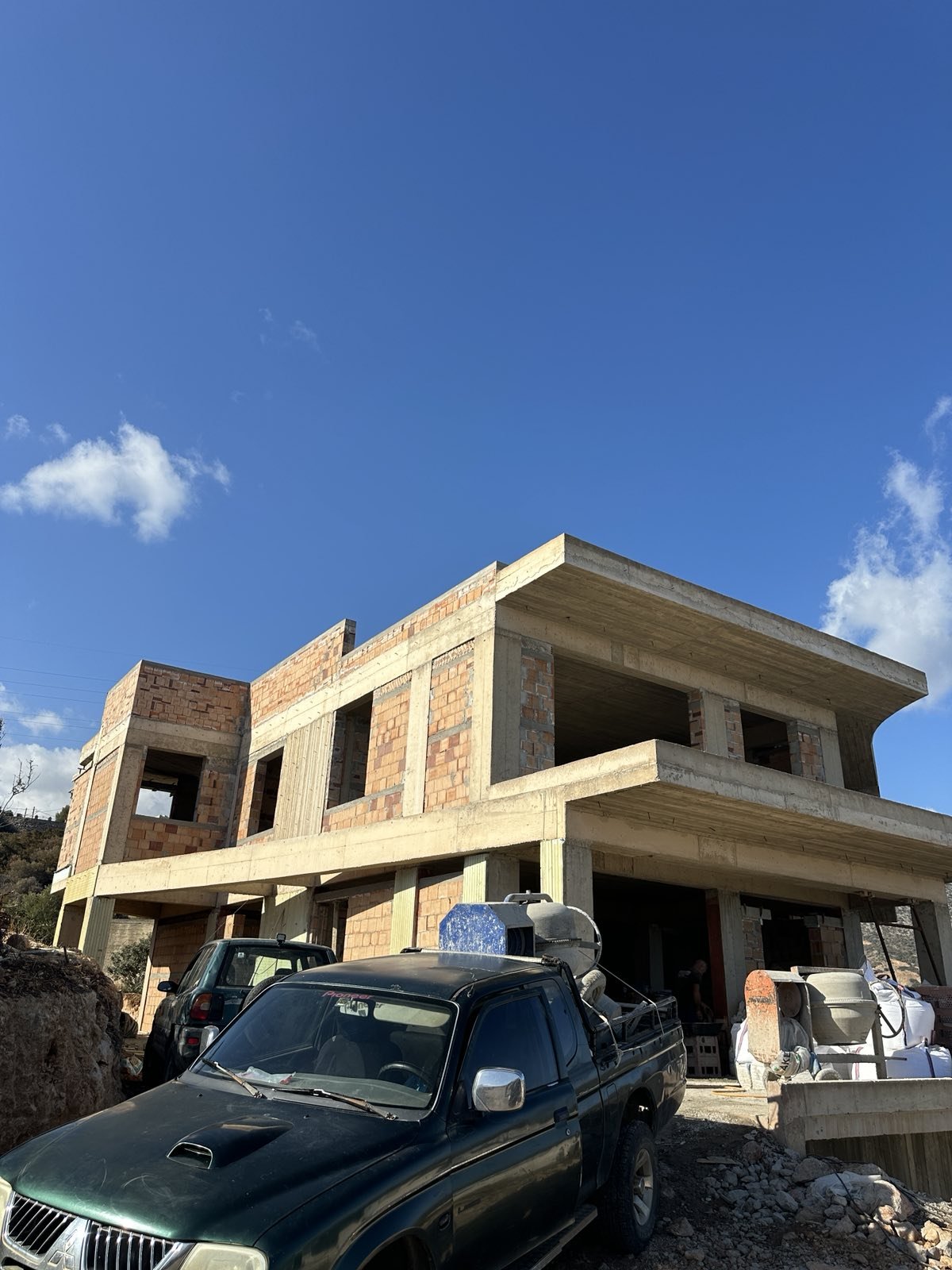 A two-story house under construction made of brick and concrete, with construction vehicles and supplies in the foreground, under a clear blue sky.