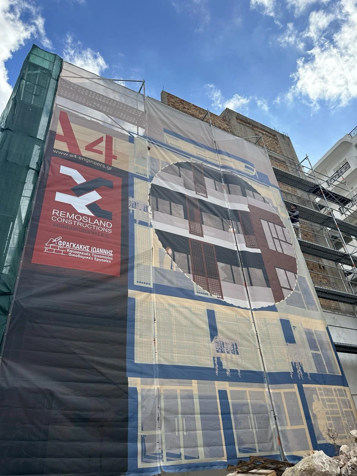 A building under construction covered by a large tarp with a printed architectural design, scaffolding on the sides, and a mostly blue sky with a few clouds above.