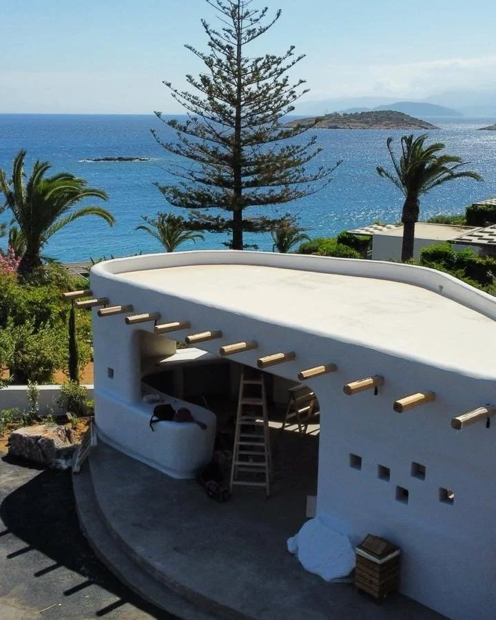 Under construction of a modern white building with curved design, overlooking the ocean with palm trees and a small island in the background.