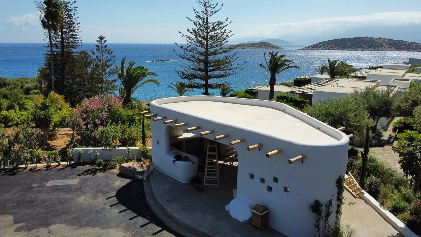A white rooftop structure under construction with wooden beams, set against a lush landscape with trees, flowering bushes, and an ocean view with islands in the background.