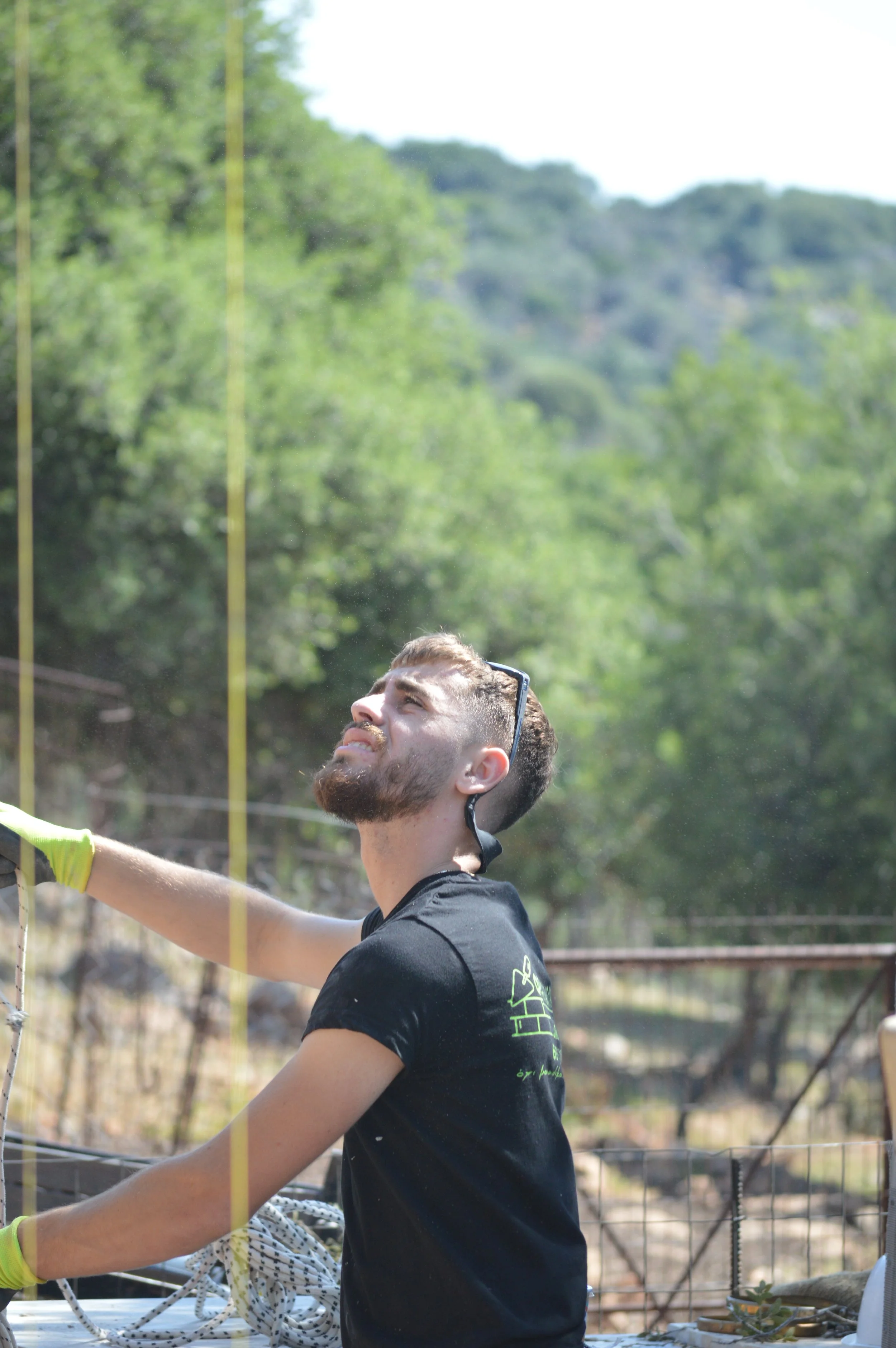 A man with a beard and short hair, wearing sunglasses on his head and a black T-shirt, stands outdoors near a fence, grimacing or looking up with a strained expression. Green trees and hills are in the background.