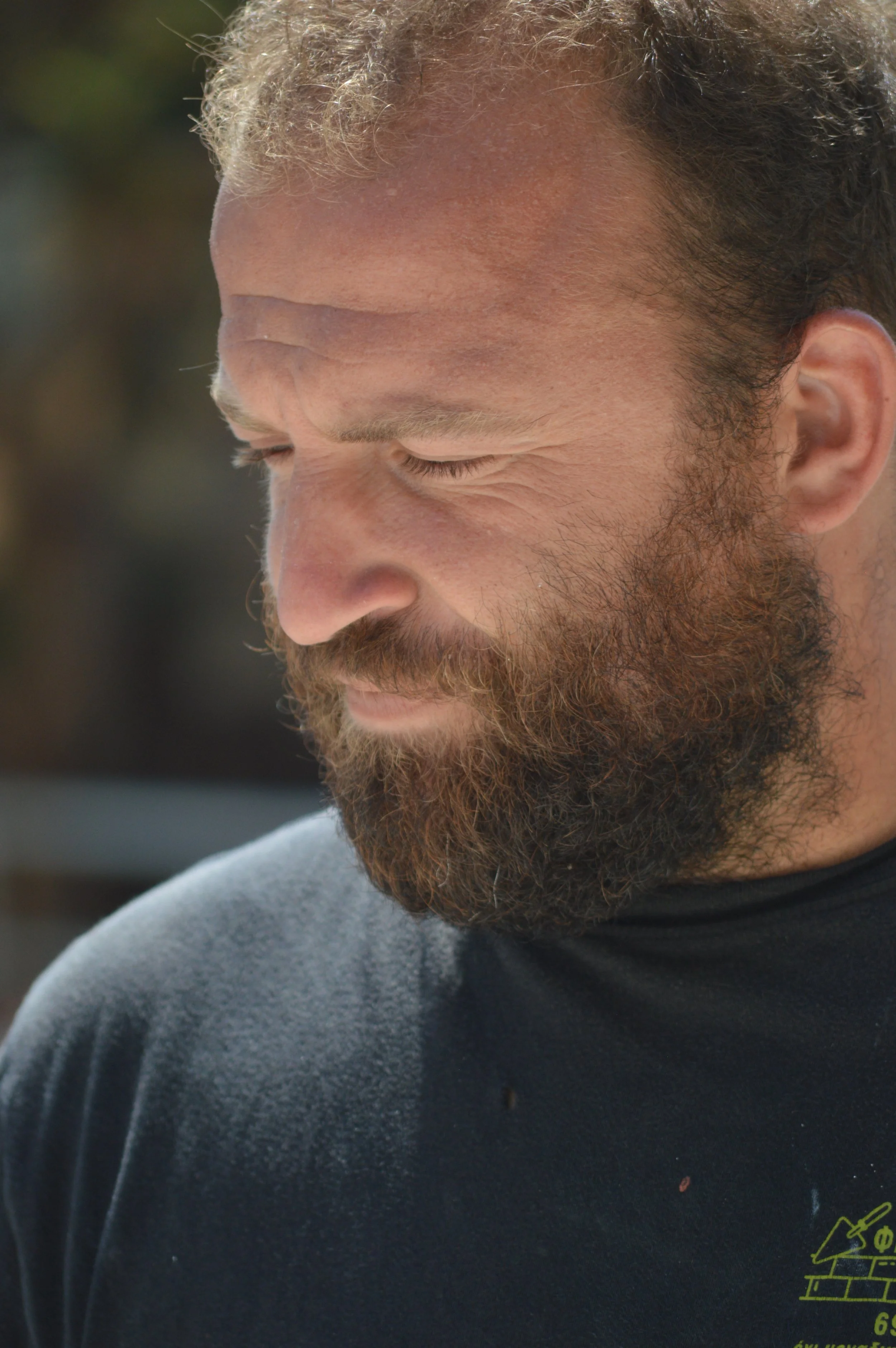 Close-up of a man with a full beard and curly hair, looking down, wearing a black shirt, outdoors.