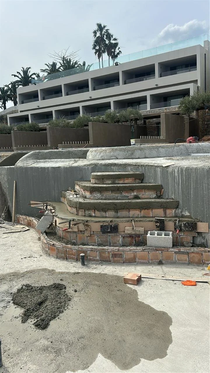 Construction site with brick stairs and unfinished concrete around a modern building with balconies, palm trees in the background, and construction materials on the ground.