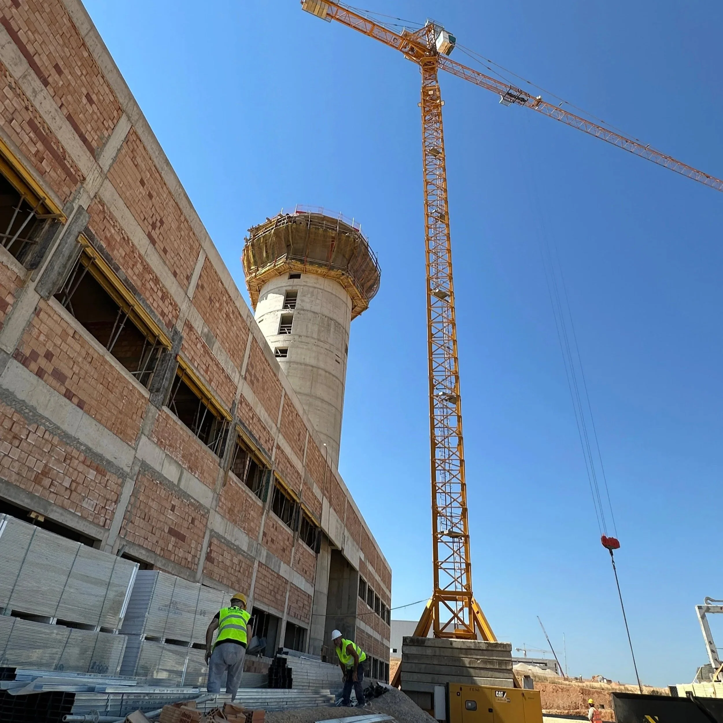 Construction site with workers, a large tower crane, and  new airport of Heraklion, Crete under construction with a concrete core and brick exterior.