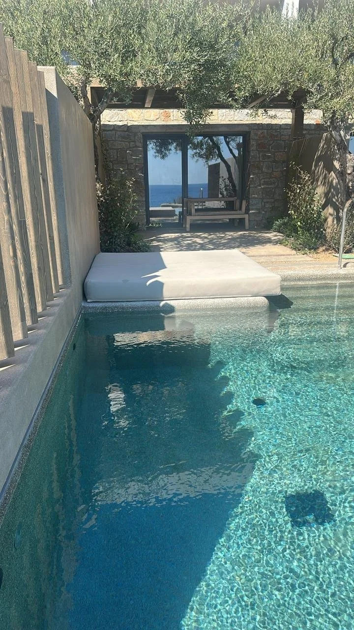 A backyard swimming pool with a white cushioned lounge chair partially in the shade, a stone wall with a window, a bench outside the window, trees, and an ocean in the background.