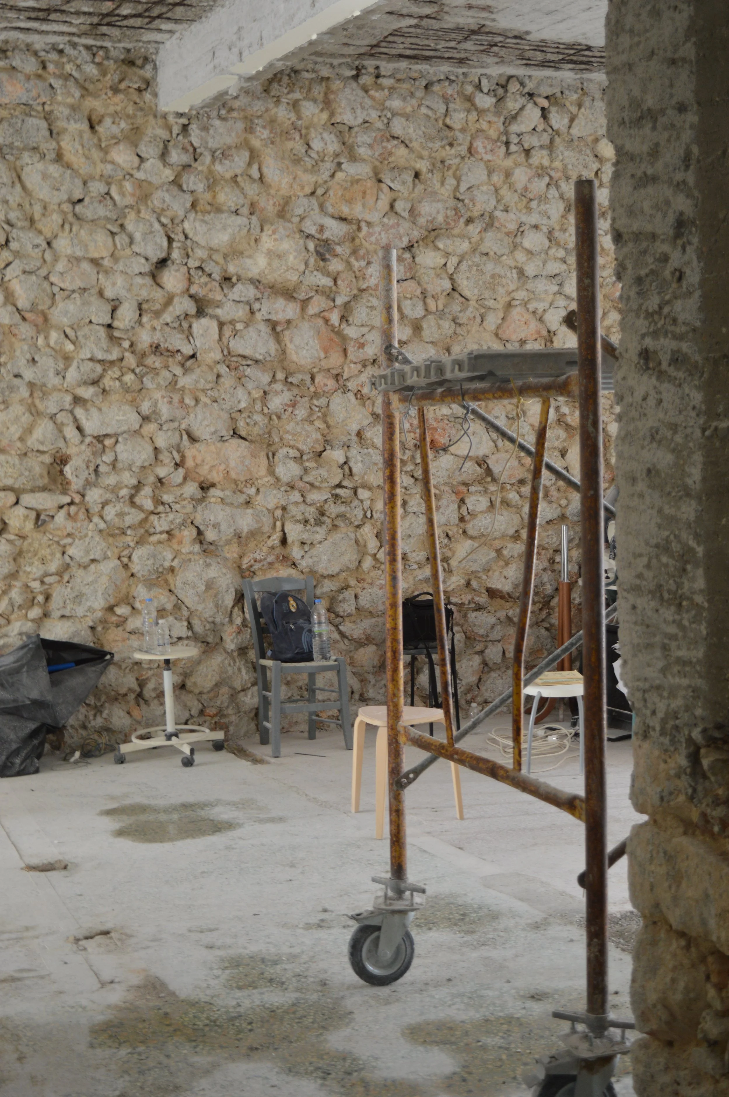 Interior of a construction site with a stone wall, metal scaffolding on wheels, two chairs, and scattered water bottles.