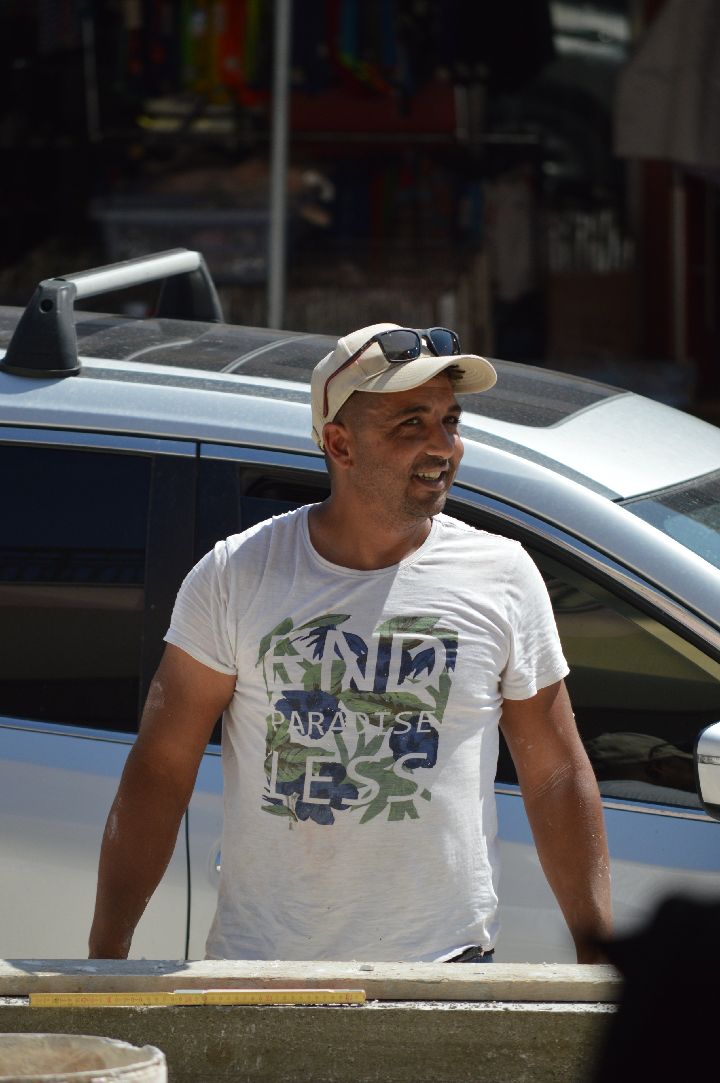 A man wearing a white cap and sunglasses on his head, smiling, standing outdoors near a silver and black vehicle, with a measuring tape on a surface in front of him.