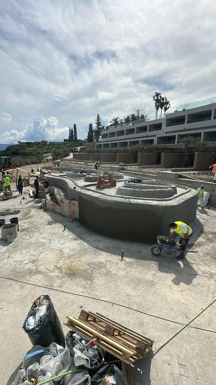 Construction site with workers building a skateboard park, with a modern building and palm trees in the background.