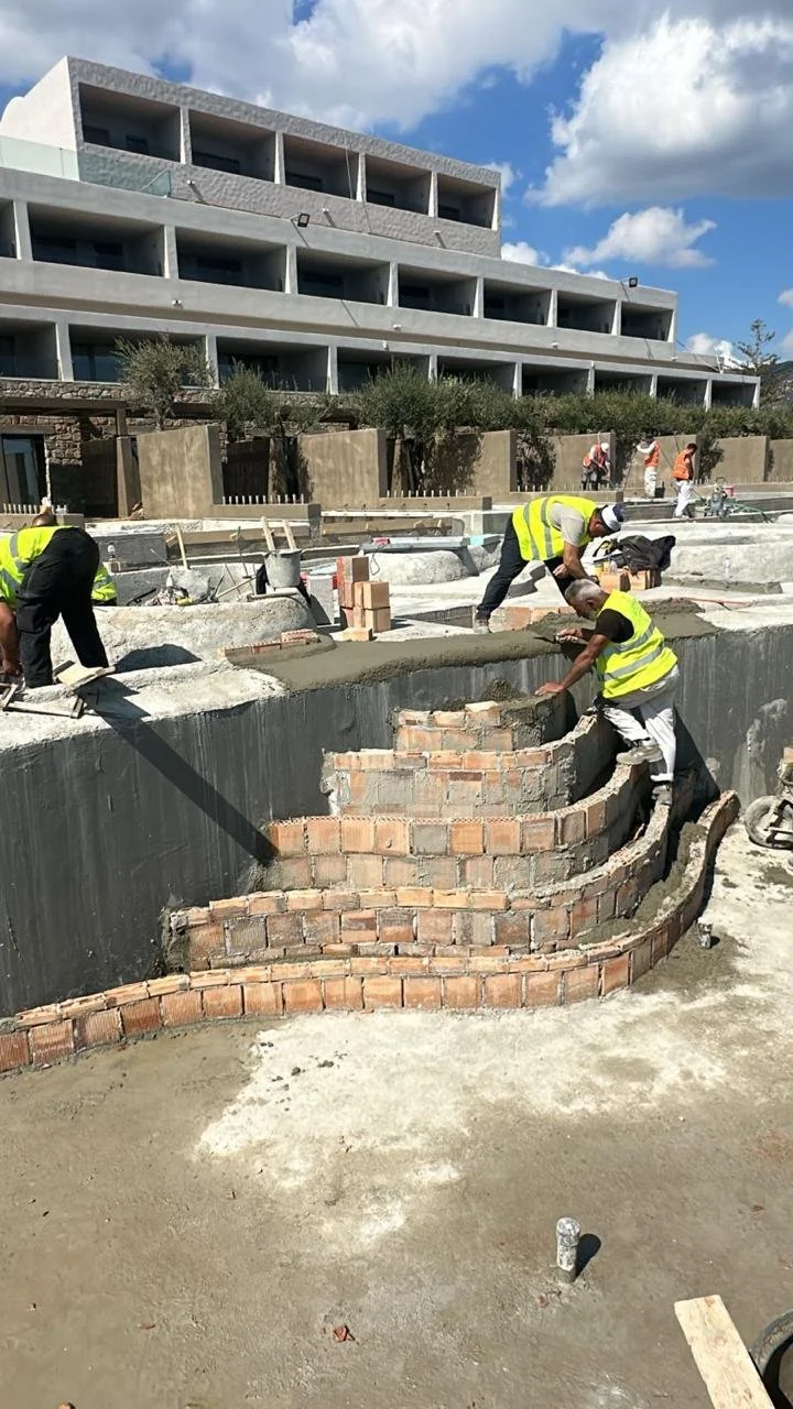 Construction workers in yellow safety vests building a brick wall around a pool or water feature at a construction site.