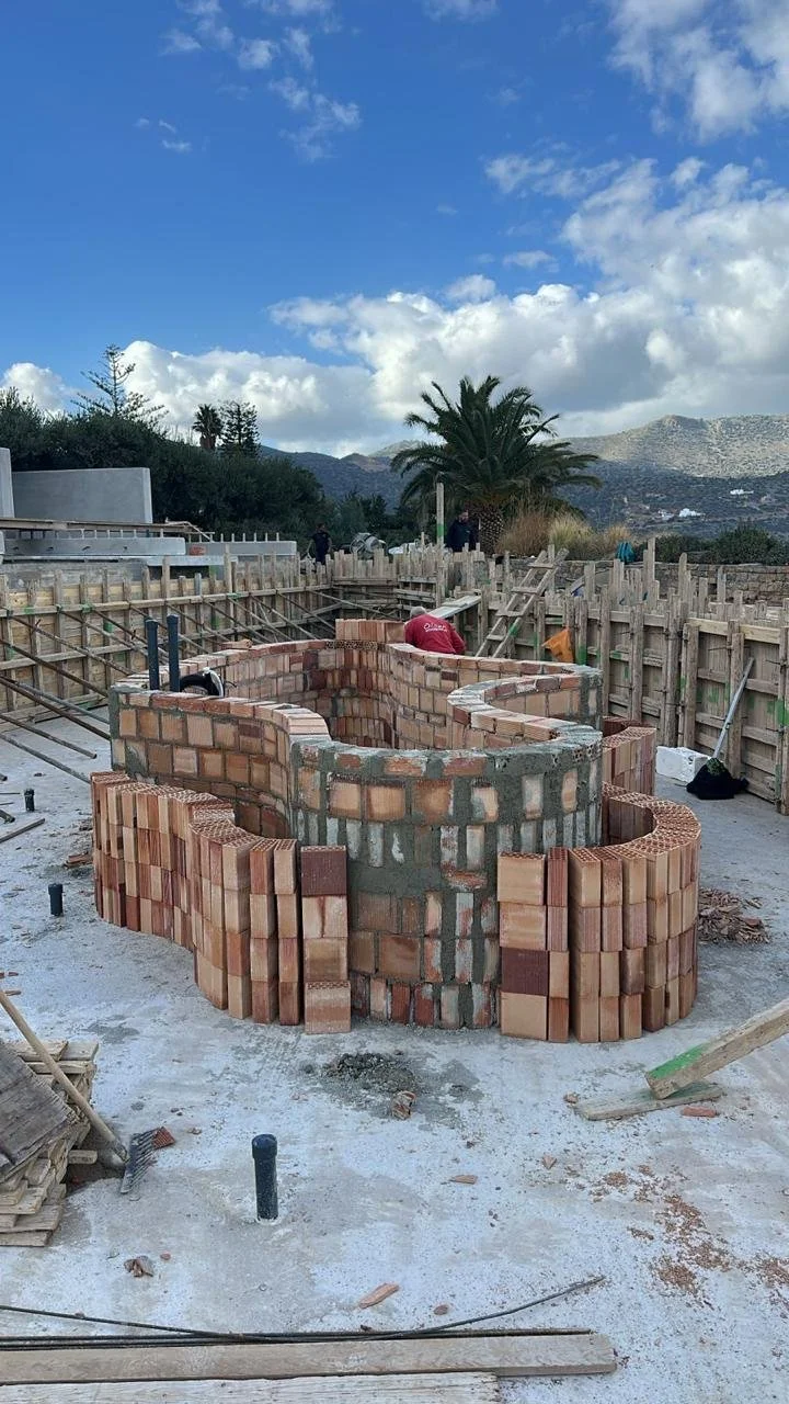 Under construction brick structure with curved walls, surrounded by wooden fencing, outdoors on a sunny day with clouds and mountains in the background.