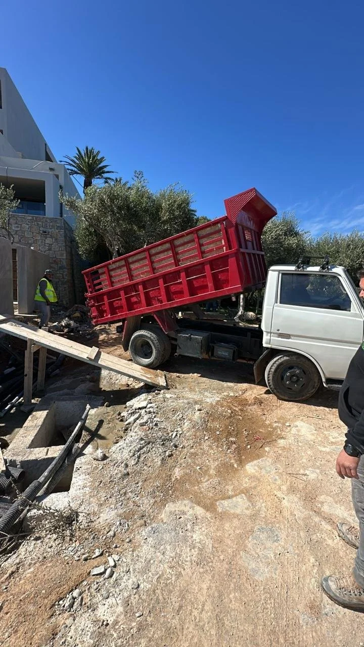 A white truck with a red cargo bed on a construction site, with the bed tilted upward. Two workers are visible, one wearing a yellow safety vest and helmet, and the other partly visible on the right side. The site has loose dirt, rocks, and construct