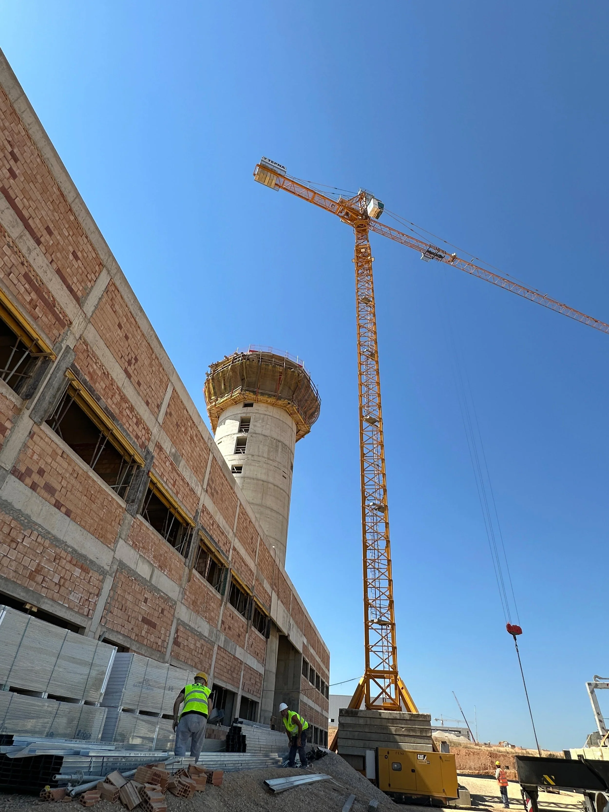 Construction site with workers, a tall crane, and new airport of Heraklion, Crete under construction, with a blue sky in the background.