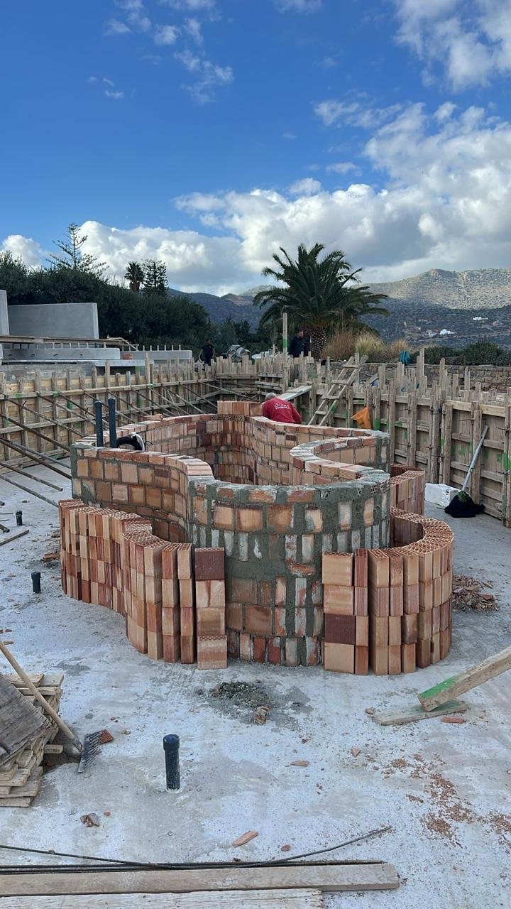 Construction site with a brick structure in progress, surrounded by wooden fencing and construction tools, with mountains and a partly cloudy sky in the background.
