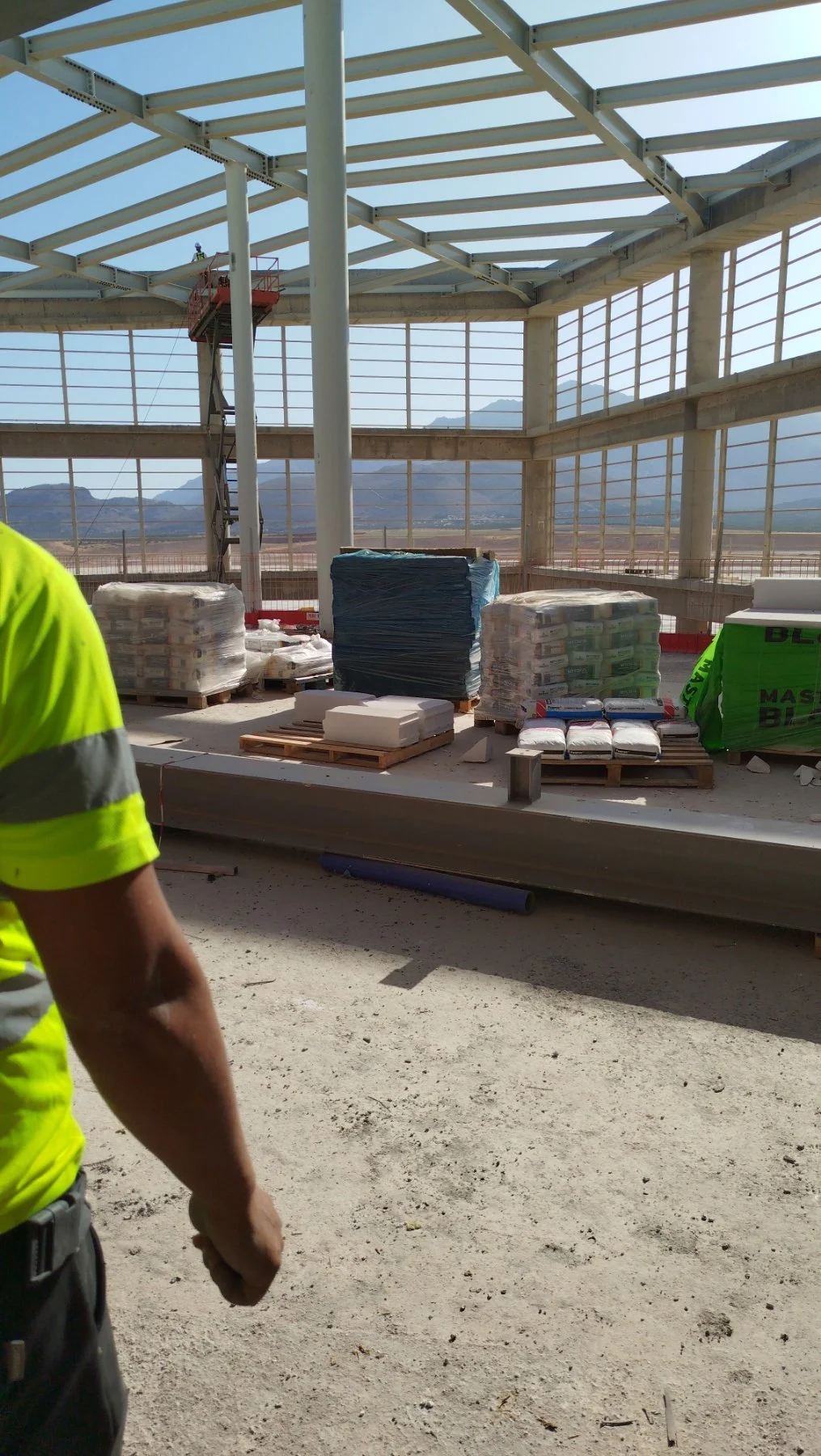 Construction site interior with building framework and pallets of construction materials, including insulation panels, cement bags, and sacks, with mountain landscape visible through large windows.