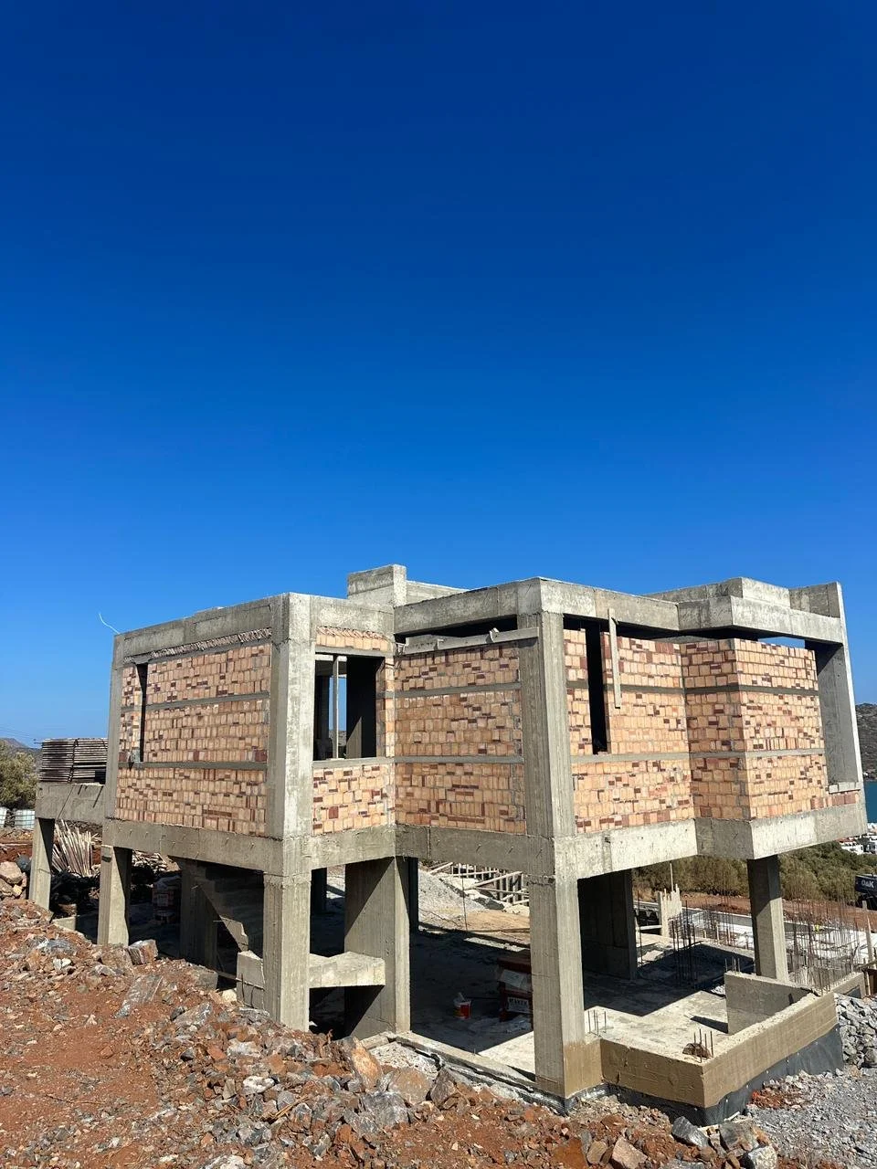Under-construction concrete building with brick walls, elevated on concrete pillars, against a clear blue sky.