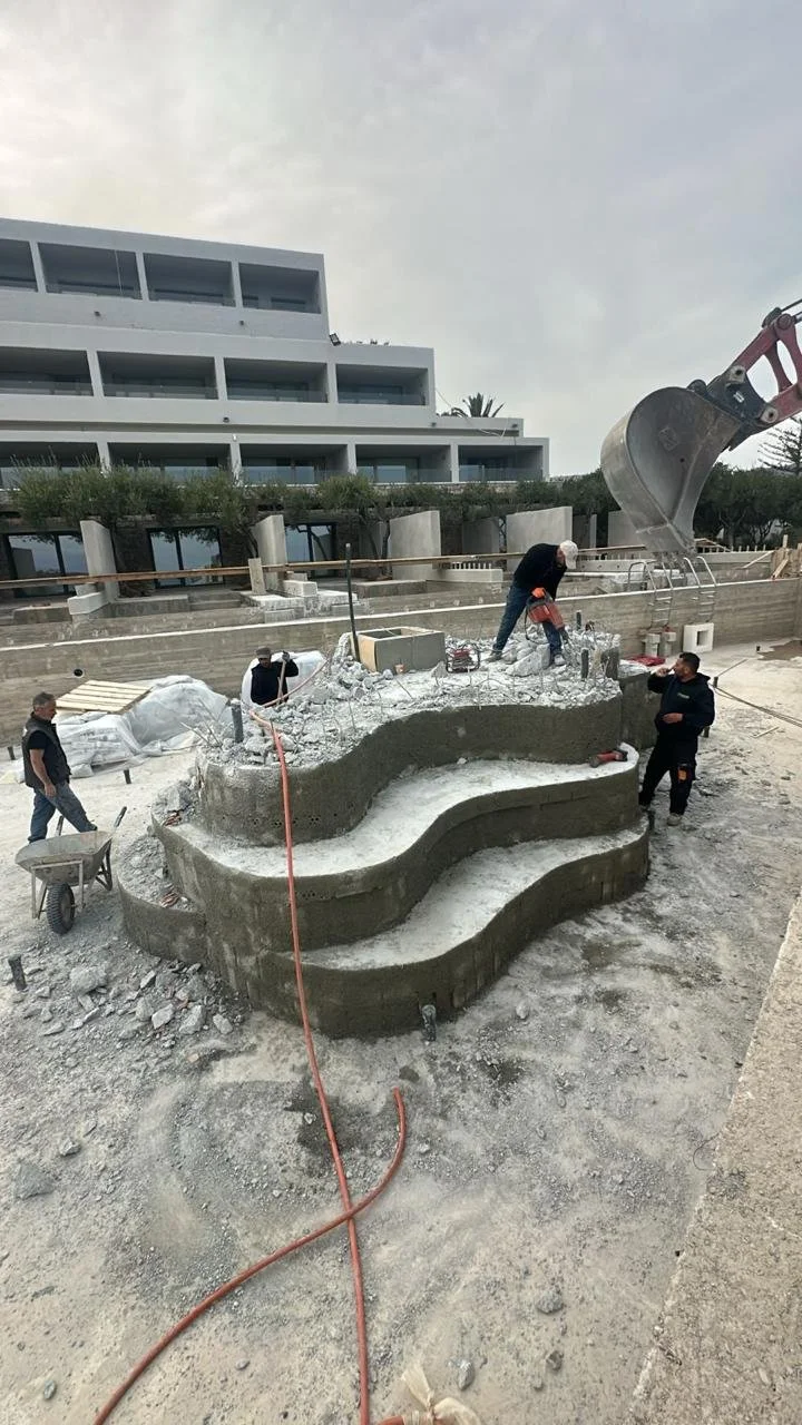 Construction workers building a multi-level outdoor structure that appears to be a stepped seating area or planter, with a large excavator in the background and a modern multi-story building behind them.