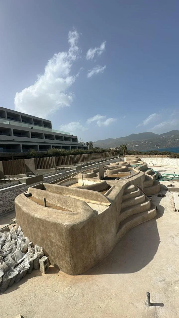 Construction site with large concrete pool shells and a modern building in the background, mountains and blue sky with clouds.