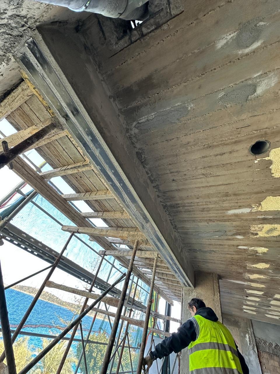 A construction worker in a neon safety vest working on scaffolding outside a building, the building's concrete ceiling and window frame are visible.