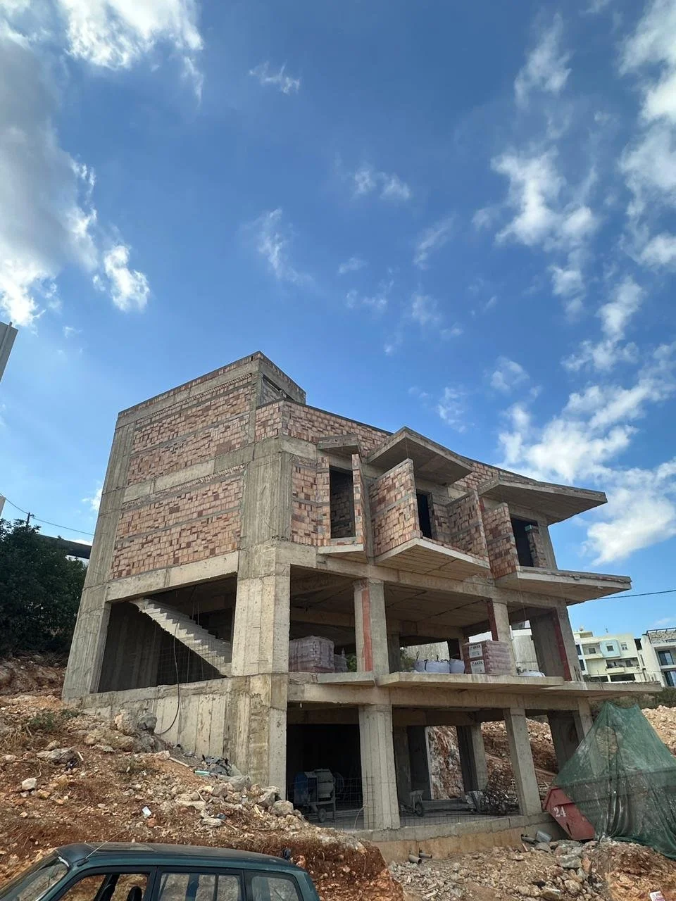 Under construction multi-story building with concrete and brick walls, exposed scaffolding, and construction materials on site under a partly cloudy sky.