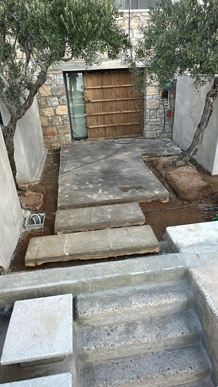 Pathway with concrete stepping stones leading to a small backyard or patio area, flanked by two trees, with a brick wall background and a wooden gate.